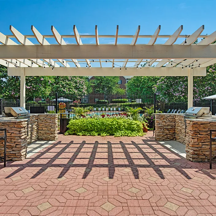 Pergola with barbecue stations and seating, surrounded by greenery and community pool view.